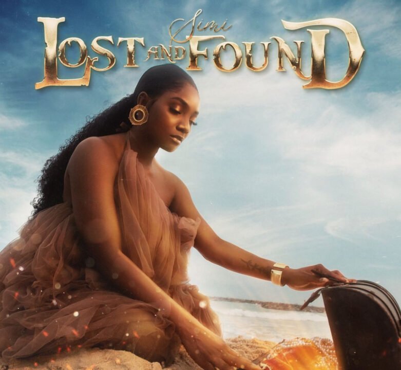 The image is a cover art for a song titled Lost and Found by Simi. It features Simi sitting on a beach, dressed in a flowing, sheer, brown gown. She is looking down and gently touching a treasure chest that is partially buried in the sand, glowing with a golden light. The background is a serene, cloudy sky with the ocean in the distance. The title "Lost and Found" is written in large, golden letters at the top, with "Simi" in a smaller, cursive font above it.